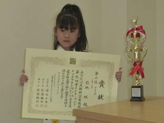 道着を着た女子生徒が立って、賞状をこちらに向けており、右の机にトロフィーが置いてある写真