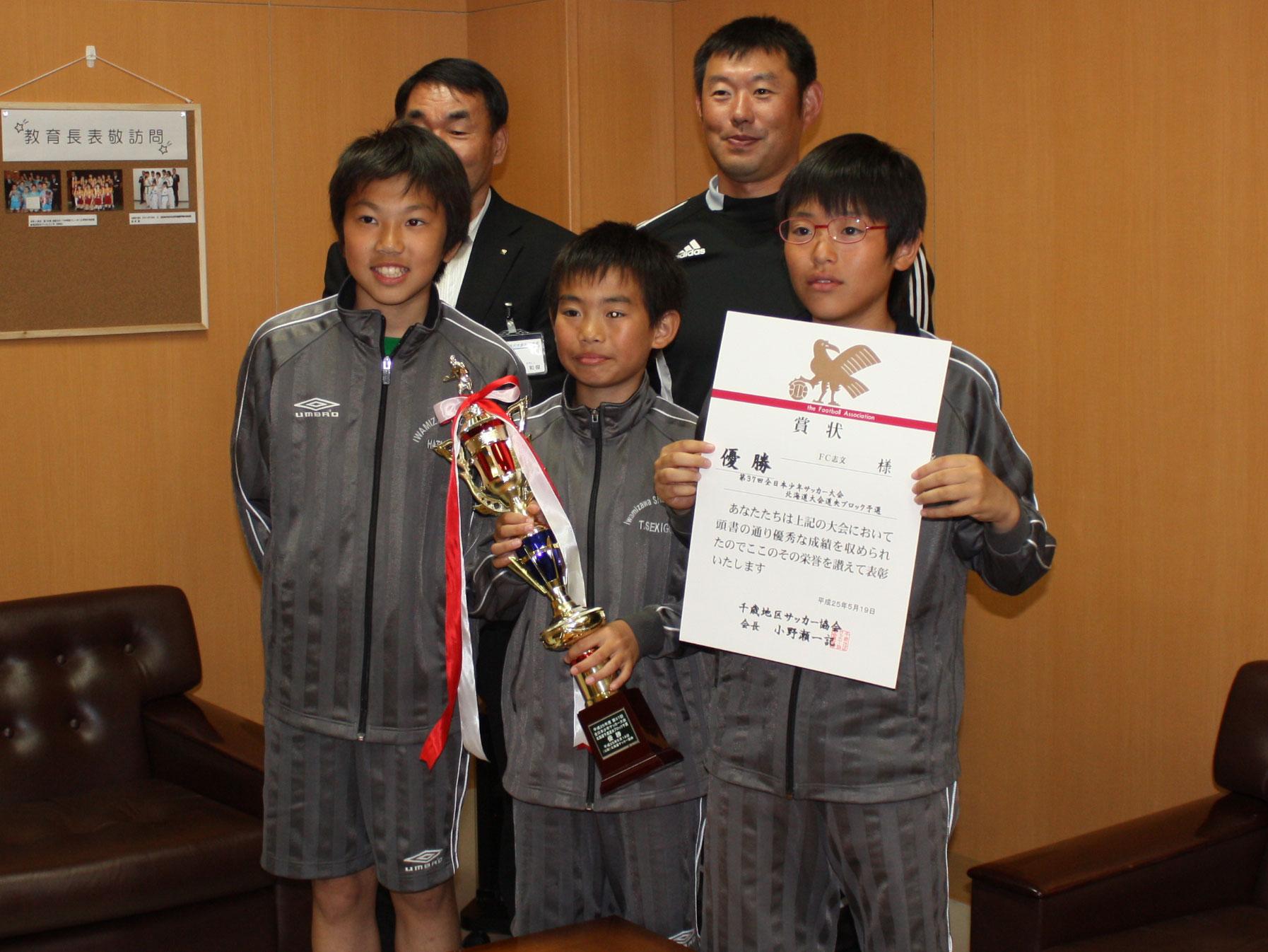 3人のユニフォームを着た男子生徒が立って、賞状とトロフィーをこちらに向けており、奥に男性2人が立って並んでいる写真