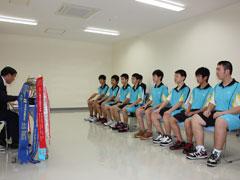 ユニフォームを着た8人の男子生徒と、その奥に1人の男性、机を挟んでスーツを着た男性が椅子に座っている写真