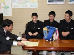 机に置かれたトロフィーをはさみ話す教育長と男子生徒の写真