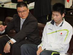 奥にスーツ姿の男性が座り、手前に白と黒と黄色のジャージを着た選手が座っている写真