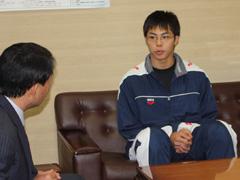 茶色いソファの上に座りながらスーツ姿の男性と話している紺と白のジャージ姿の選手の写真
