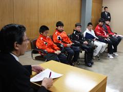 スーツを着た男性の前に座るジャージを着た児童たちの写真