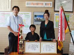 二枚の賞状とトロフィーや旗と一緒に記念撮影をする小学生生徒の写真