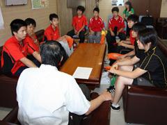 ソファーに座りユニフォーム姿で教育長の話を聞くバドミントン選手の学生たちの写真