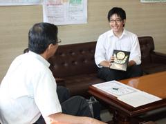 室内で賞状を持った制服姿の学生が教育長と対談している写真