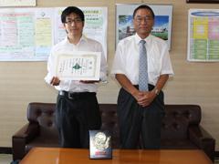 室内で賞状を持った制服姿の学生が教育長と並んで撮影されている写真