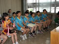 青いユニフォーム姿の小学生男子と黄色と赤のユニフォーム姿の小学生女子達が椅子に座って教育長と対面している様子の写真