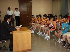 青いユニフォーム姿の小学生男子と黄色と赤のユニフォーム姿の小学生女子達が椅子に座り、教育長が子どもたちへ奨励している様子の写真