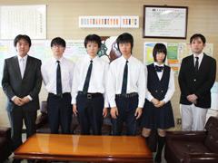 室内にて制服姿の学生達が顧問と並んで撮影されている写真