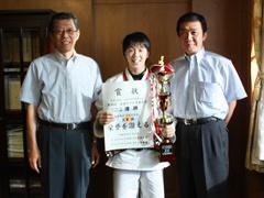 右から、ワイシャツを着た男性、道着を着て賞状とトロフィーをこちらに向けている女子生徒、ワイシャツを着た男性が並んで立っている写真