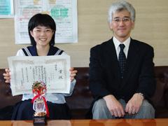 右からスーツを着た男性と、制服を着て賞状をこちらに向けている女子生徒が椅子に座っていて、女子生徒の前にはトロフィーが置いてある写真