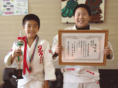 右から白い道着を着て賞状をこちらに向けている男子生徒と、白い道着を着てトロフィーををこちらに向けている男子生徒が並んで立っている写真