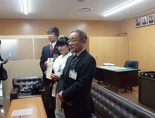 スーツを着た教育長と男性の間に道着姿の女子生徒が賞状を持って立っている様子を横から撮った写真