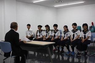 スーツを着た男性と机を挟んで、6人の白い制服を着た女子生徒と、1人の白い制服を着た男子生徒とが椅子に座っている写真