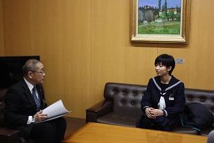 木壁と絵画が飾ってある室内で教育長と学生服の女学生が黒いソファーに机を挟んで座り対談している写真