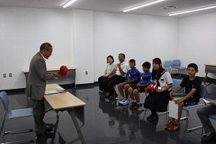 スーツを着た男性と机を挟んで、4人の生徒と、1人の男性と、1人の女性が椅子に座っている画像