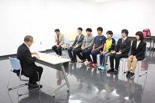 スーツを着た男性と机を挟んで、7人の方が並んで座っている写真
