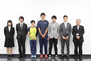 右から、スーツを着た男性が2人と、灰色のジャケットを着ている男性と、青い服を着ている男性と、黄色い服をを着ている男性と、スーツを着ている男性と、スーツを着ている女性が並んで立っている写真