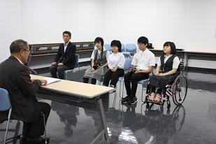 スーツを着た男性と机を挟んで、制服を着た車椅子に座っている女子生徒と、制服を着た2人の男子生徒と、制服を着た女子生徒と、スーツを着た男性が椅子に座っている写真
