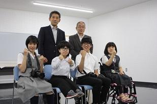右から、制服を着た車椅子に座っている女子生徒と、制服を着た2人の男子生徒と、制服を着た女子生徒が座り、後ろにスーツを着た2人の男性が並んでいる写真