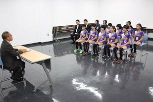 スーツを着た男性と机を挟んで、複数のユニフォームを着た女子生徒と、複数のスーツを着た方が座っている写真