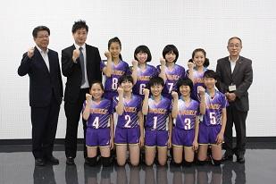 右から、スーツを着た男性と、複数のユニフォームを着た女子生徒と、2人のスーツを着た男性が並んでいる写真