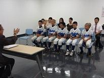 スーツを着た男性と机を挟んで、複数ののユニフォームを着た男子生徒、奥に複数の方が椅子に座っている画像