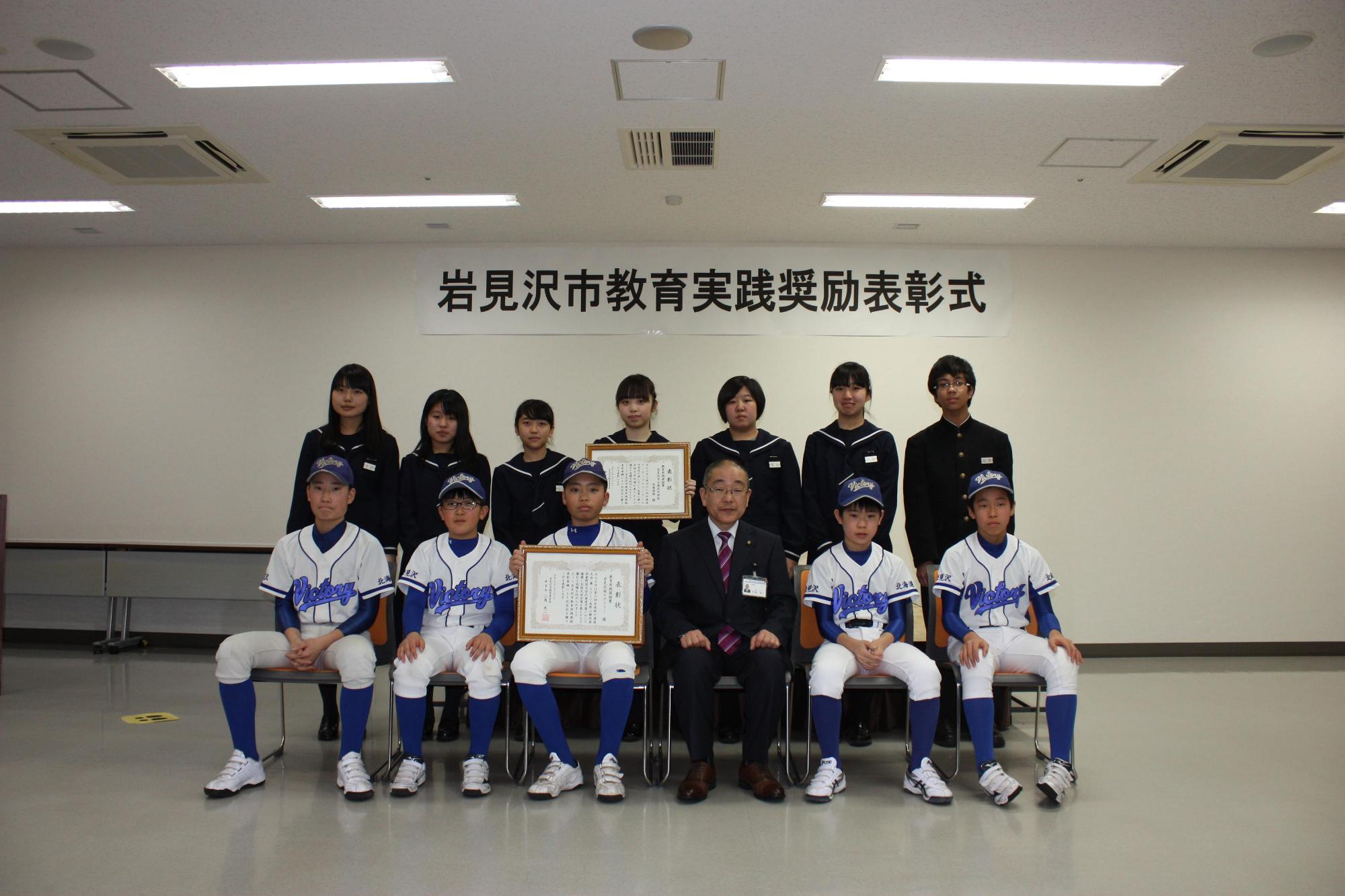8名の受賞者が手に賞状を持ち座っていて前列には青いユニフォームを着て立っている1団体の受賞者たちの写真