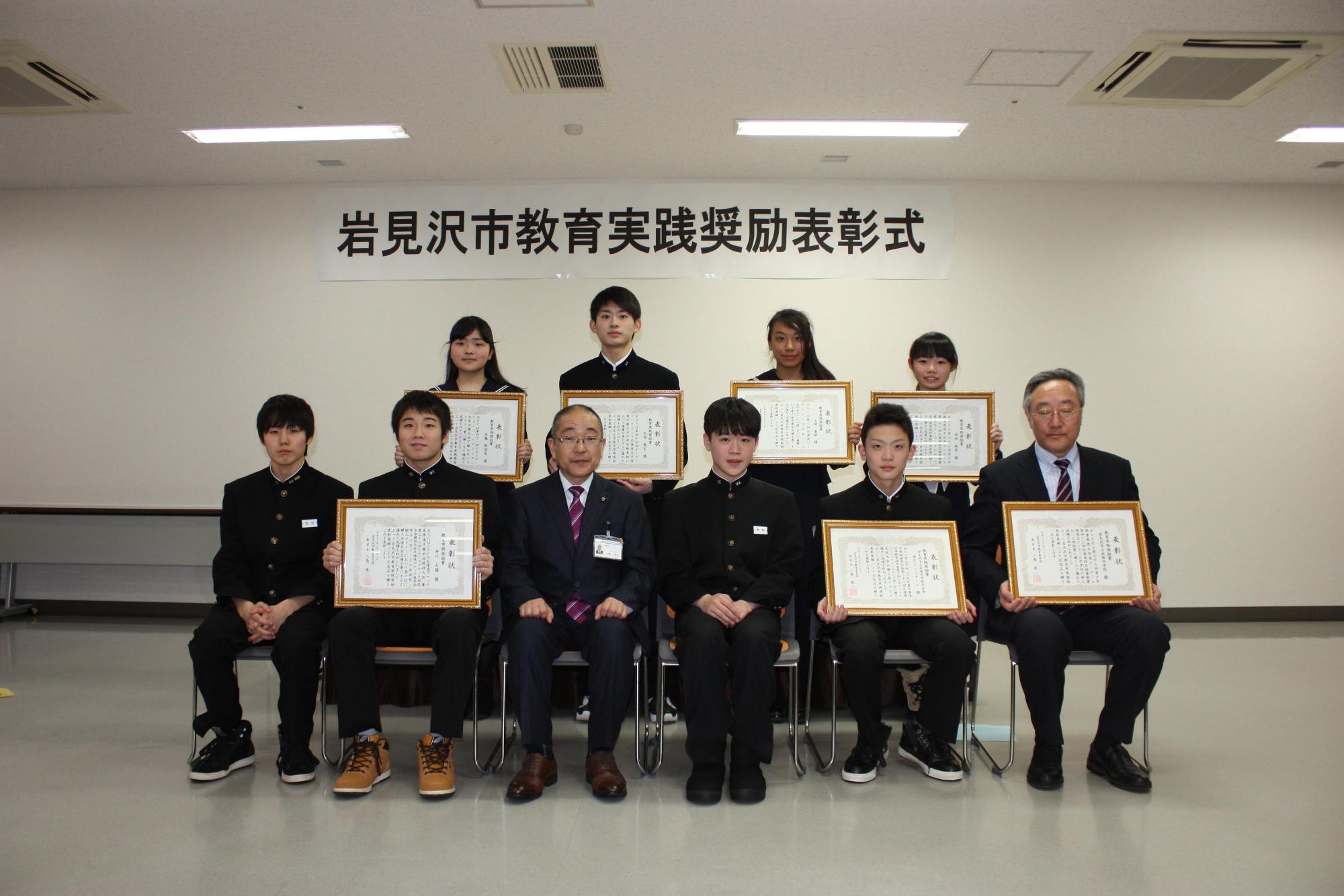 賞状を持つ7名の人と何も手にせず座っている人の写真