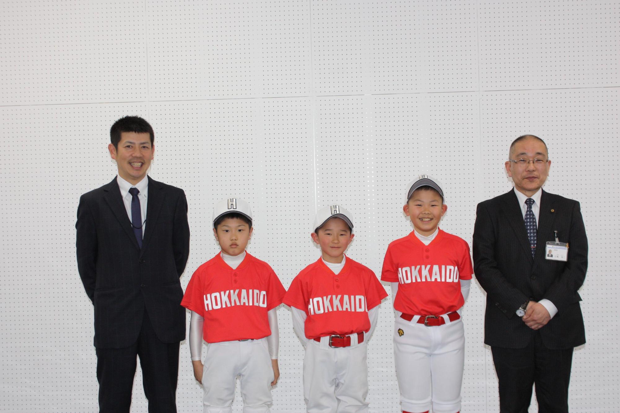 軟式野球大会に出場するユニフォーム姿の児童と教育長とスーツの男性が横一列に並んだ写真