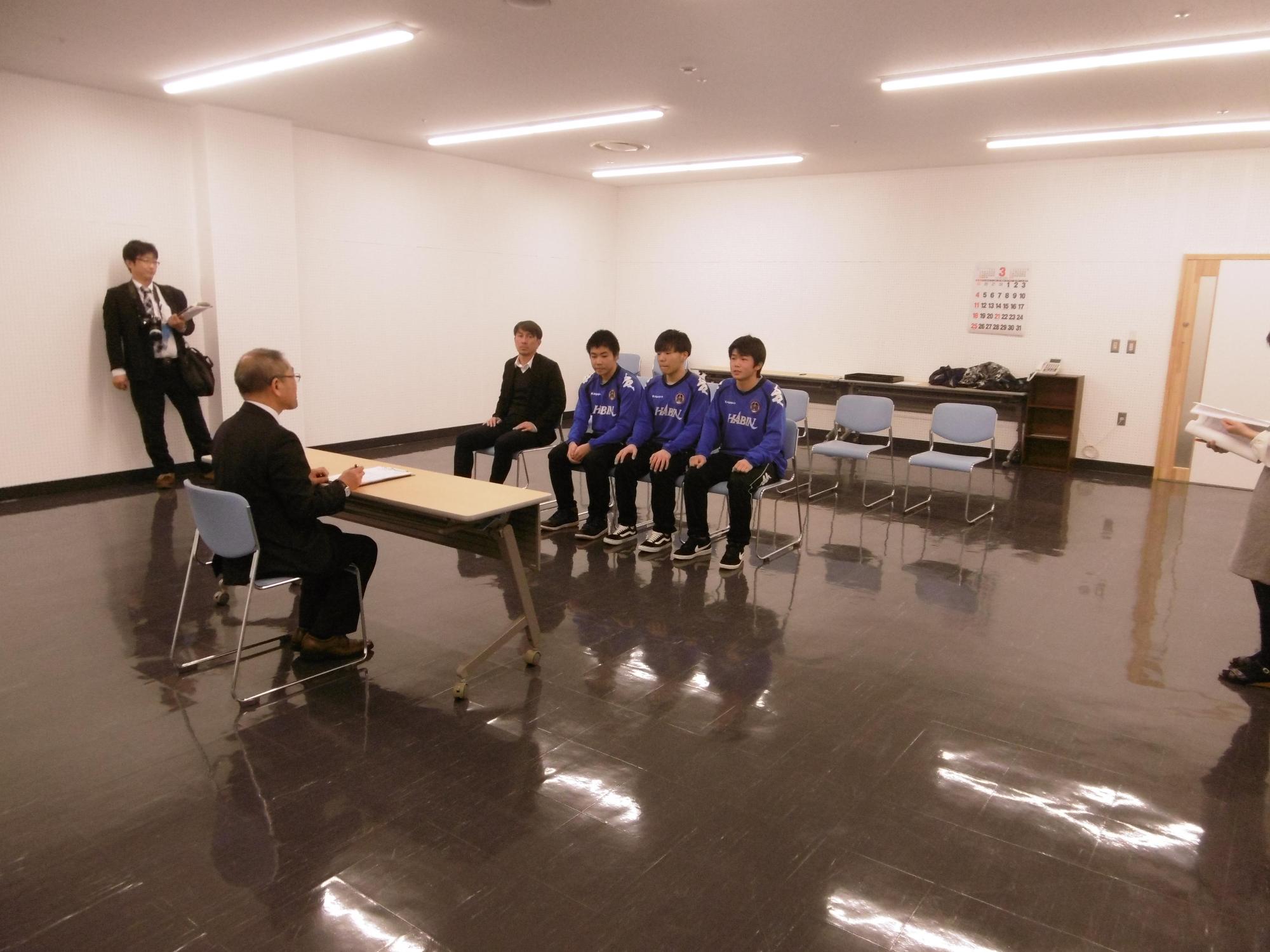 フットサル全国大会に出場した学生と教育長とスーツの男性が対面して座る写真