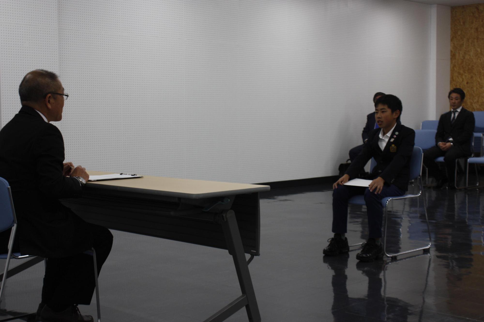 白壁に囲まれた室内で市長と制服の小学生が机向かいに談義している写真