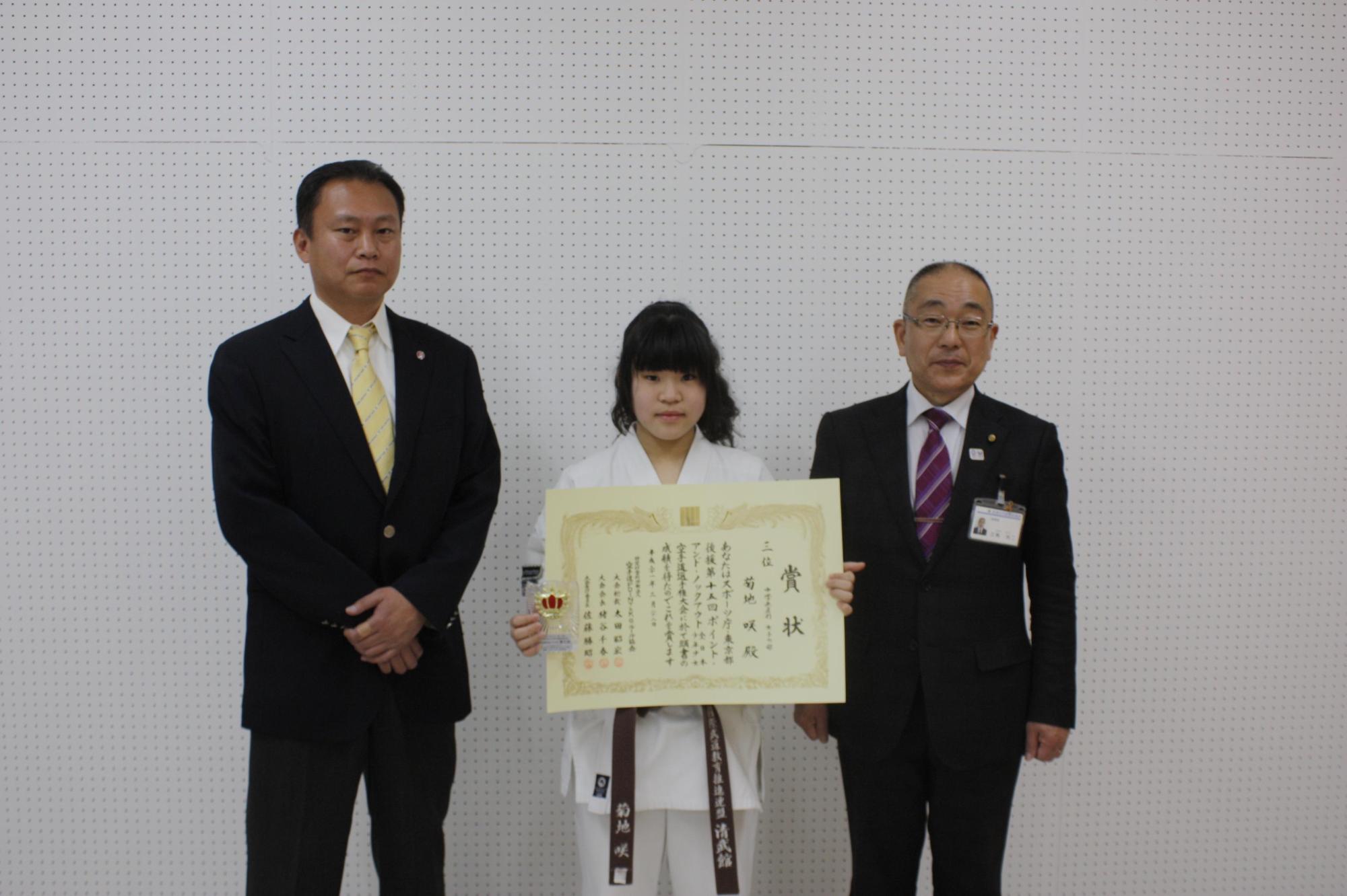 表彰状を両手で持った菊地 咲さんを囲む教育長の男性と教育委員会の男性が3人で並んでいる写真