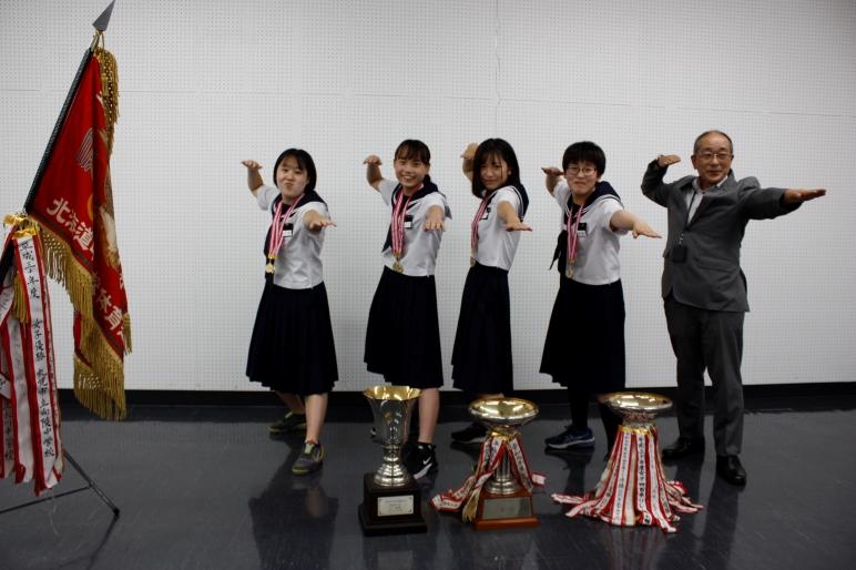 首にメダルをかけた女子生徒が教育長と水をかくようなポーズで優勝カップの前で撮影している写真