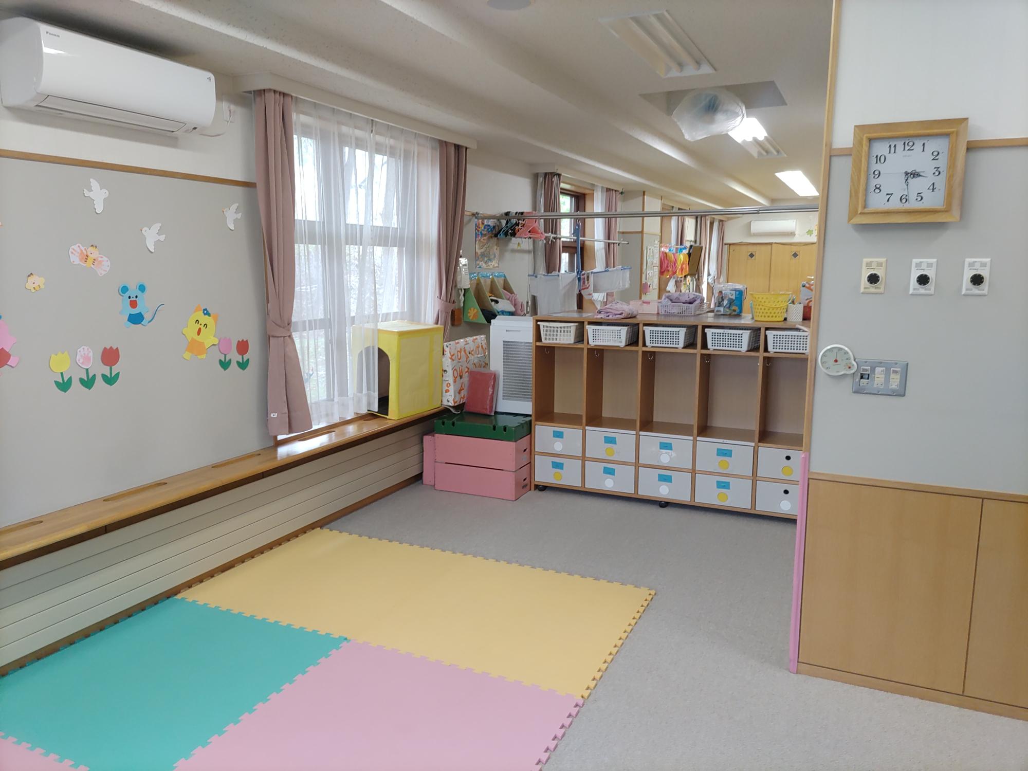 白いカーペットの上に色とりどりのマットが敷いてあり、右の壁に木枠の時計がかけてある未満児（0歳児）保育室その2