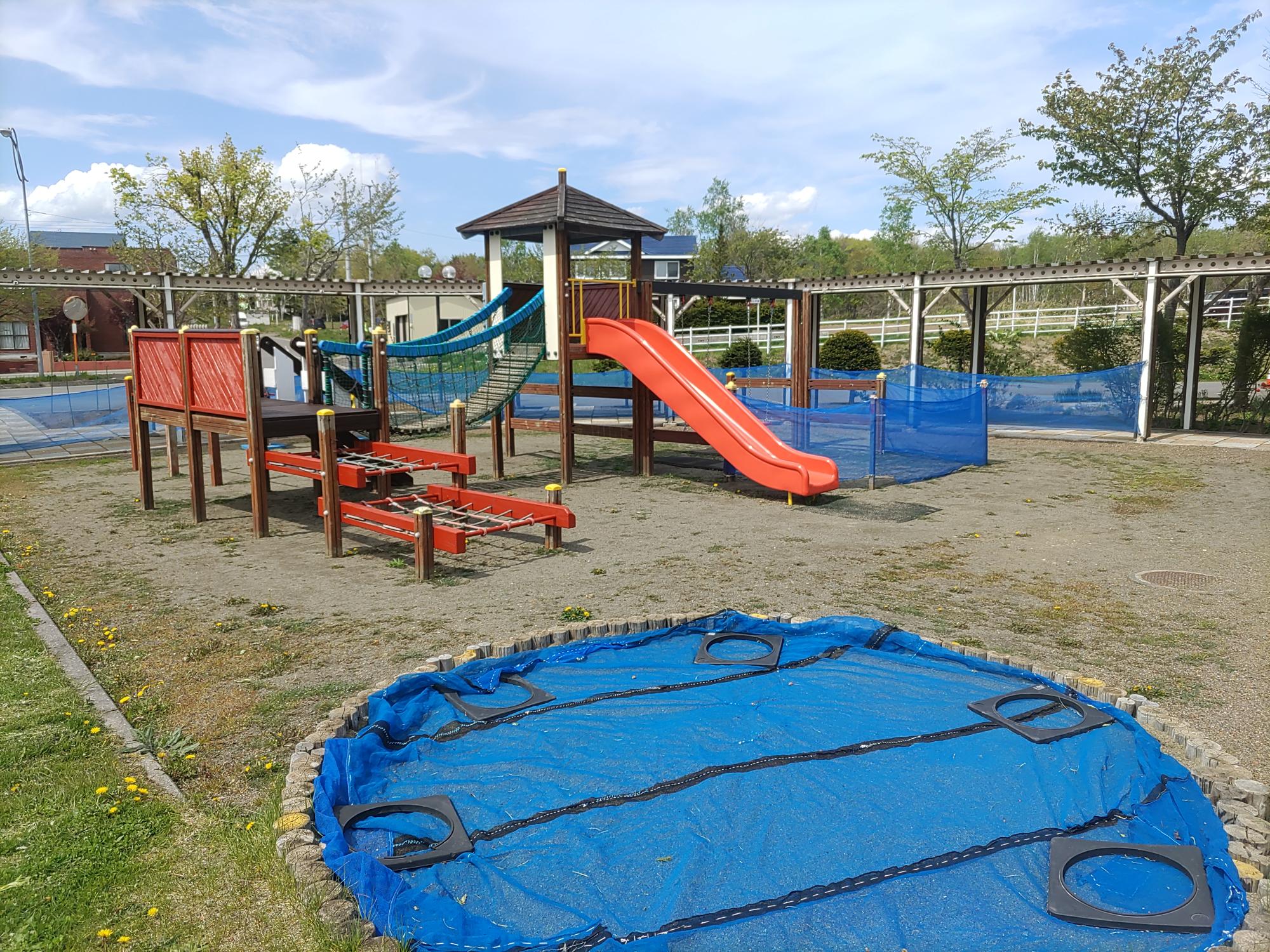 奥に滑り台や吊り橋などのある複合遊具があり、手前にブルーシートがかけられた砂場が写る園庭の写真