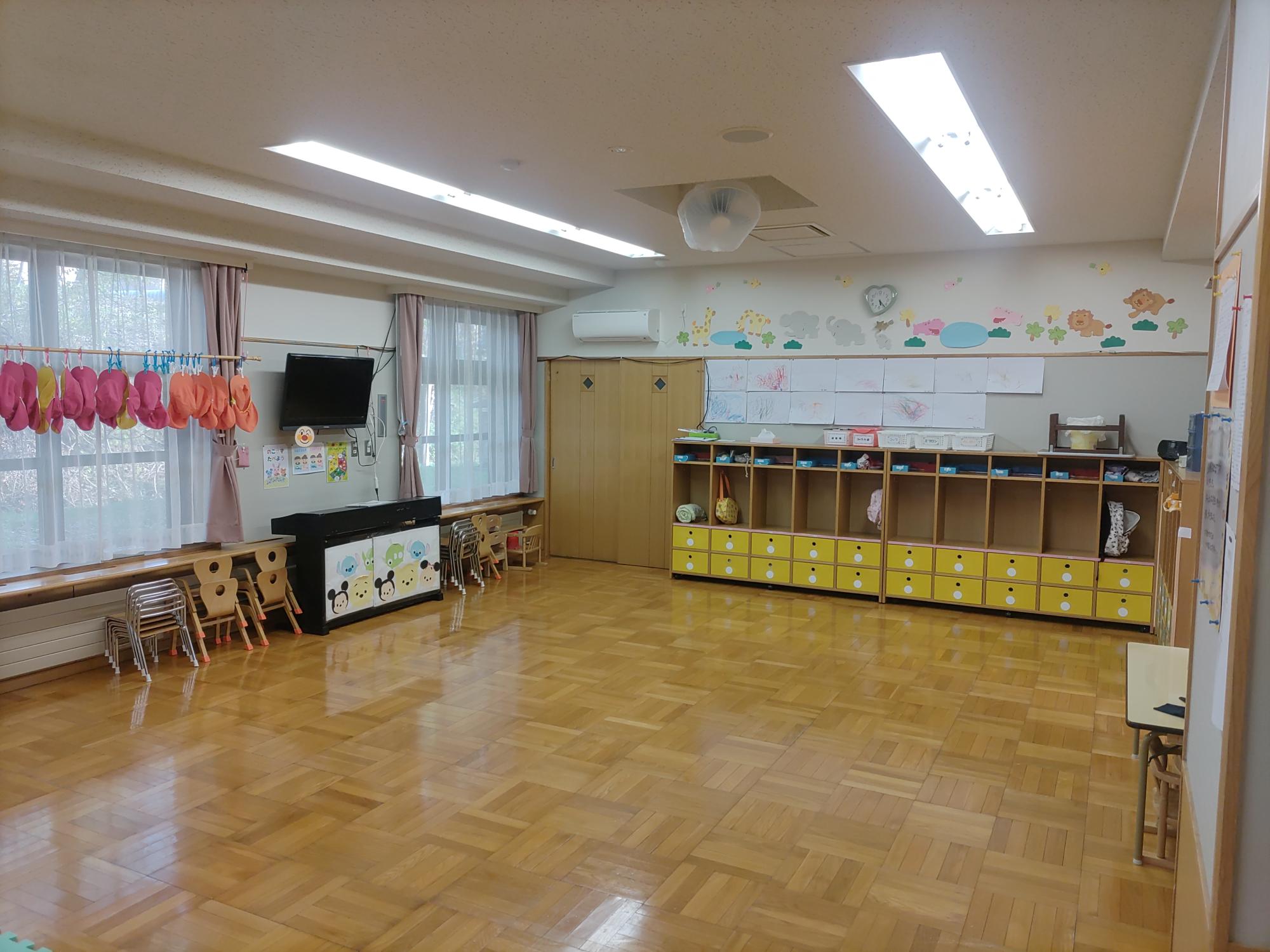 フローリングの床に白い壁、奥に棚があり、左の窓側に体育帽子がつるされている未満児（1、2歳児）保育室