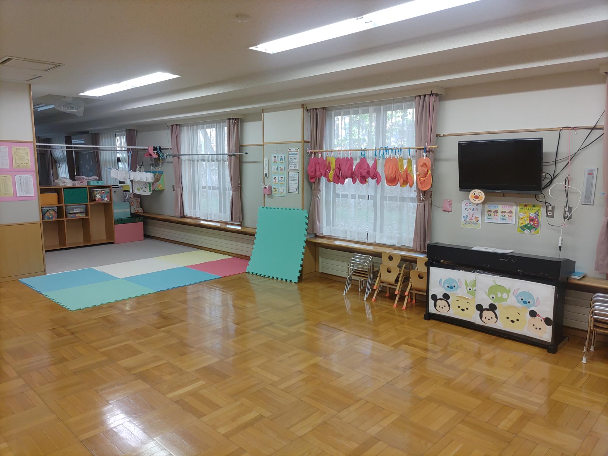 フローリングの床に白い壁、奥に色とりどりのマットを敷いたプレイルームがある未満児（1、2歳児）保育室その2