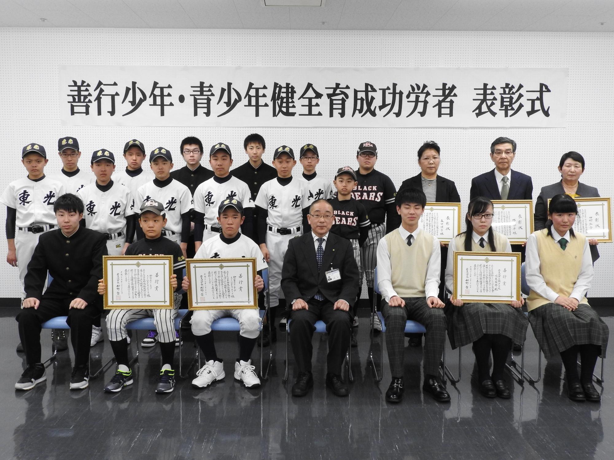善行少年・青少年健全育成功労者表彰式の横断幕の前に並ぶ、表彰状を持った少年少女と大人たちの写真