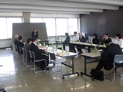 四角形に並べられた机の前の椅子に座り会議に参加する人々と、奥の机の前に立ちマイクを持ち話すスーツ姿の男性の写真