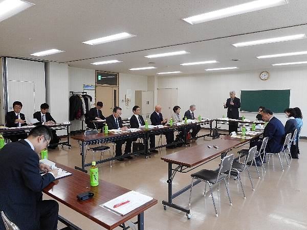 四角形に並べられた机の前に座る参加者たちと、マイクを持ち立って話すスーツ姿の男性の写真