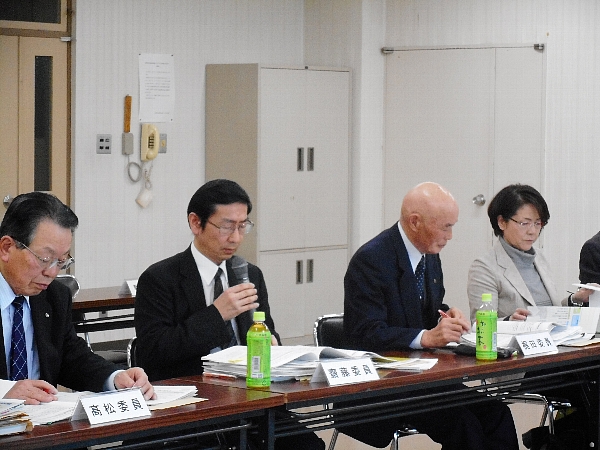 4人の参加者が机の前の椅子に座っており、そのうちの一人のスーツ姿の男性がマイクを持っている写真
