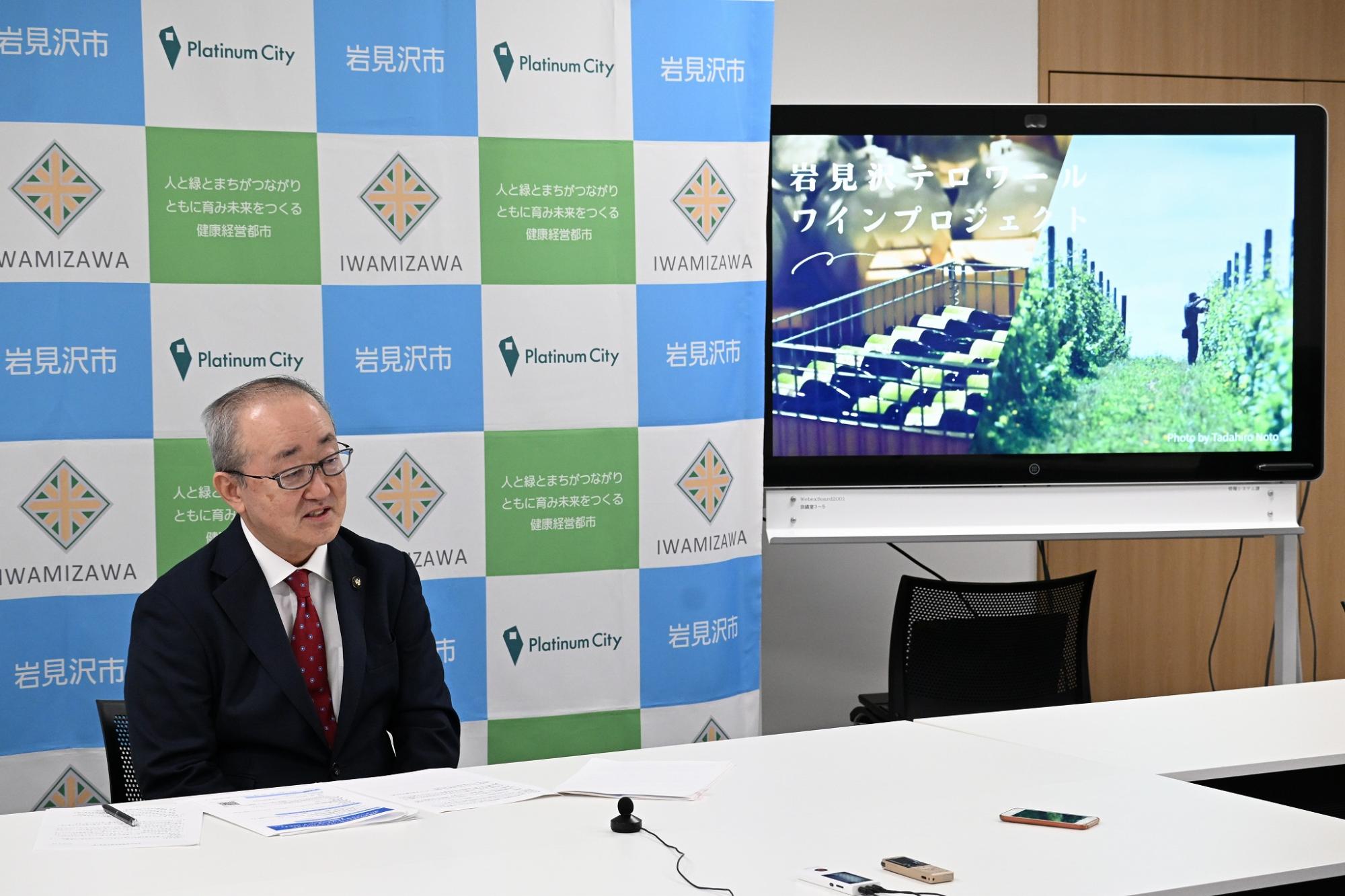 ワインとブドウ畑が映し出されたモニターの前で岩見沢テロワールワインプロジェクトについて説明する松野市長