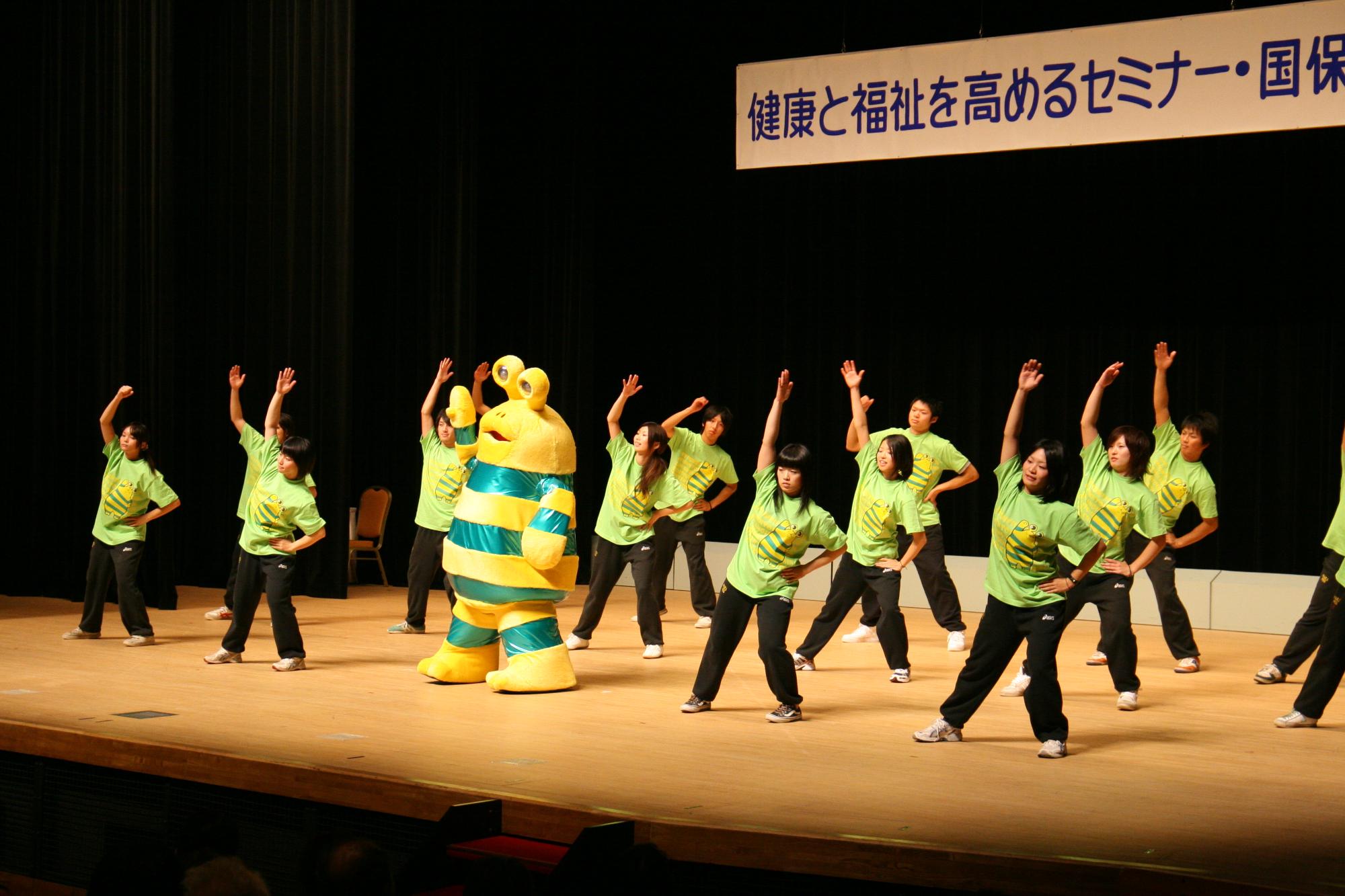 健康と福祉を高めるセミナー国保運動教室の看板が掲げられた大きな舞台の上で、そろいの緑色の服をきて、中心に黄色と緑のキャラクターが配置し運動をしているひゃっぴい体操の写真