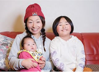 2人の子どもを抱えてソファーに座っている小林睦美さんの写真