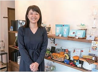 カフェ店内でコーヒー豆や小物類が並ぶ棚の前に立っている矢島幸子さんの写真