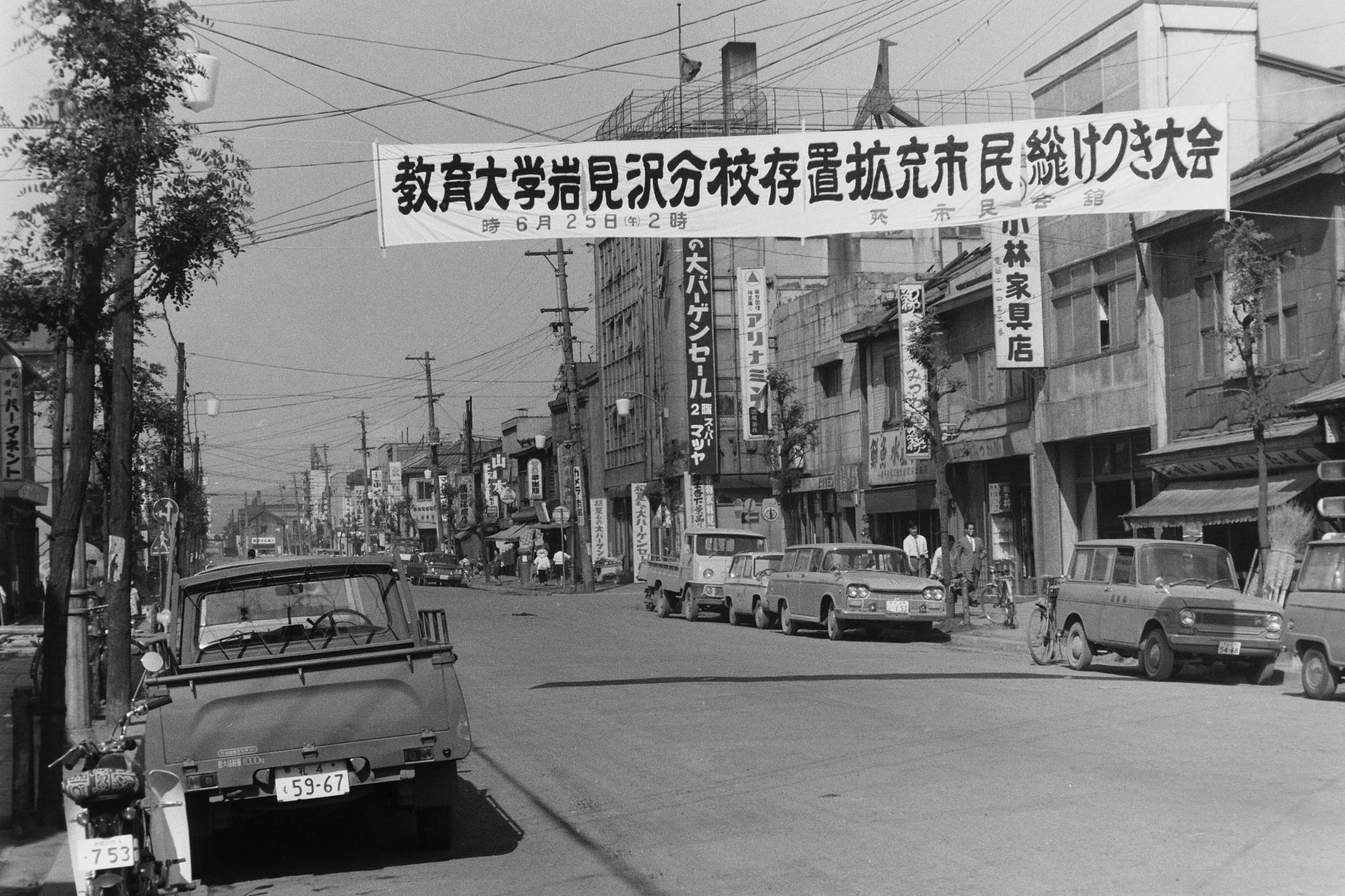 両脇に商店が並ぶ大通りの上に、教育大学岩見沢分校存置拡充市民総けつき大会の横断幕が掲げられている昭和41年の教育大学岩見沢分校の存続拡充運動の様子の白黒写真