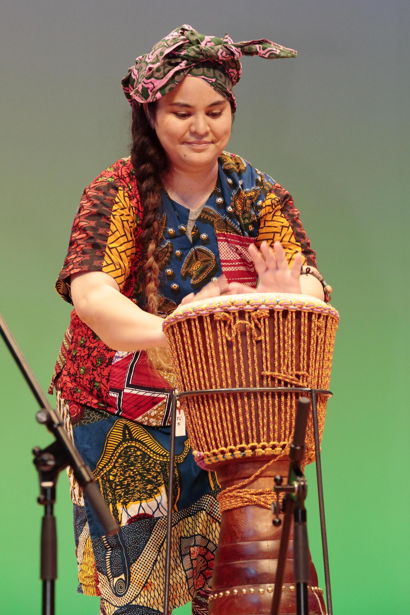 紐編みで側面を装飾された小太鼓のような形をしたアフリカの打楽器「ジャンベ」を演奏する女性の写真