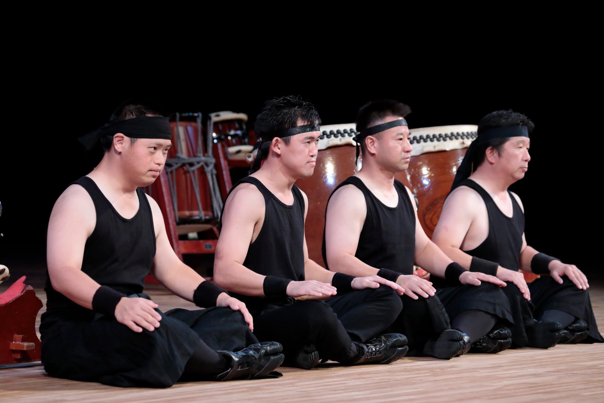 舞台の上で胡坐をかいている、黒い鉢巻を巻いて和太鼓の衣装を着た男性たちの写真